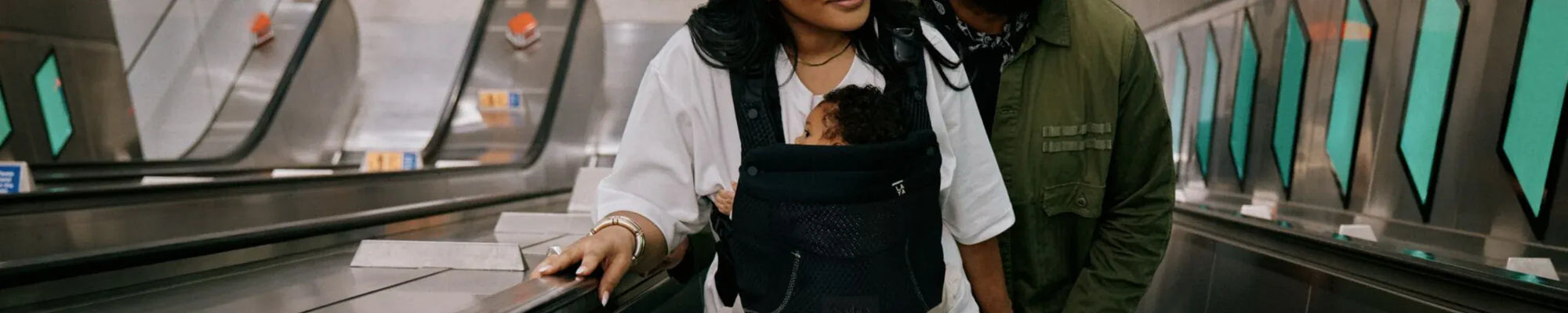 Parents using a black soft structured baby carrier on an escalator