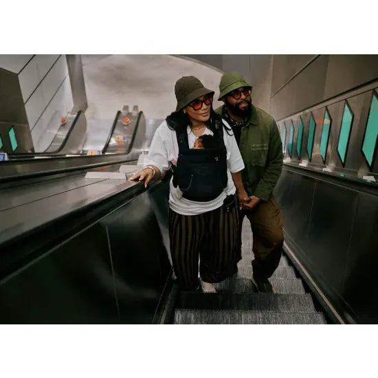 Two parents with baby carried in the Cybex Laya Baby Carrier in Black, riding an escalator in an urban transit setting