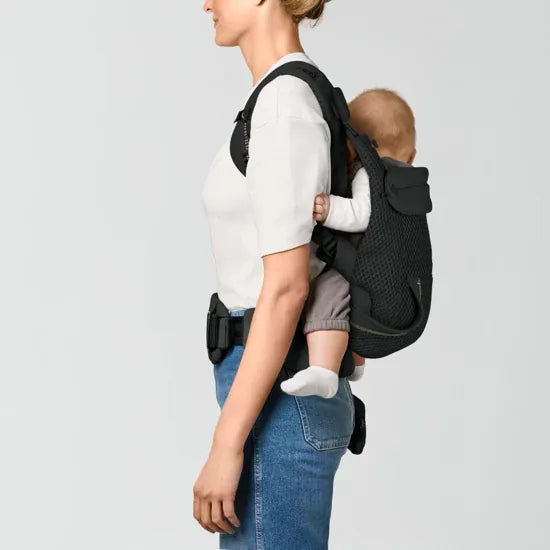 Parent carrying baby in back carry position in the Cybex Laya Baby Carrier in Black, side profile view